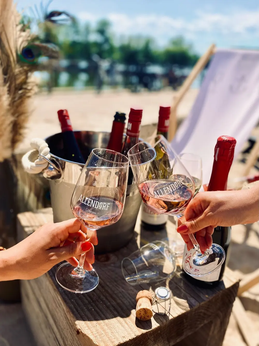 Zwei Gläser Rosé stoßen an, dahinter ein Weinkühler mit Allendorf-Flaschen und ein Liegestuhl am Strand
